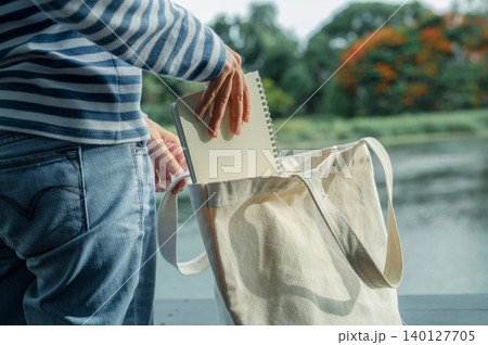 Woman placing notebook into sustainable canvas tote bag. Eco friendly fashion and mindful living. Sustainable fashion and minimalist lifestyle. Sustainable lifestyle choices. Slow life. Green habits. 140127705