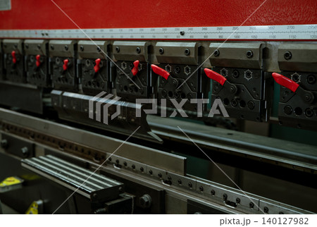 CNC metal press brake with aligned tooling and red locking handles. Operators shape steel components in an industrial workshop. Precision, controlled force and accurate setup every fabrication step. 140127982