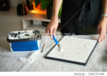 Artist making textured canvas art. Woman applying white plaster paste with a blue palette knife at table covered with protective sheet at home. Concept of handmade craft and creative hobby Artist making textured canvas art. Woman applying white plaster paste with a blue palette knife at table covered with protective sheet at home. Concept of handmade craft and creative hobby 140131164