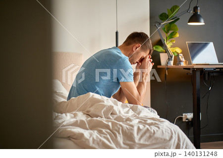 Man sitting on unmade bed with hands near face in bedroom. Concept of stress, anxiety, fatigue and mental wellbeing at home. Man sitting on unmade bed with hands near face in bedroom. Concept of stress, anxiety, fatigue and mental wellbeing at home. 140131248