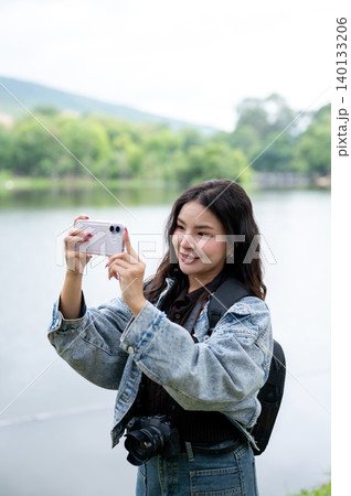 Pretty asian woman with backpack holding smartphone taking a photo beside reservoir lake in the park Pretty asian woman with backpack holding smartphone taking a photo beside reservoir lake in the park 140133206