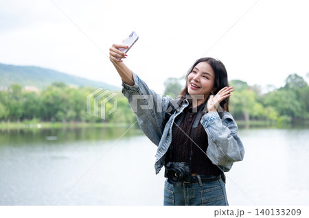 Pretty asian woman with camera using looking at smartphone as standing beside reservoir lake in park Pretty asian woman with camera using looking at smartphone as standing beside reservoir lake in park 140133209