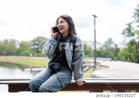 Happy asian woman in denim carrying a camera talking on phone while sitting on wooden bench in park. 140133227