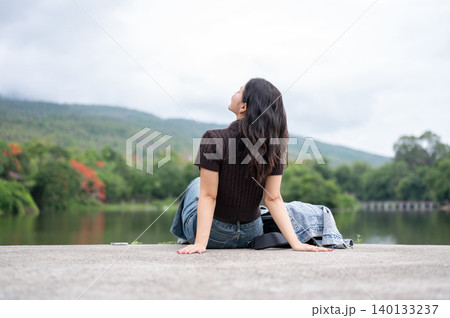 Back view of asian woman chilling beside lake across mountain while sitting on concrete road ground. Back view of asian woman chilling beside lake across mountain while sitting on concrete road ground. 140133237
