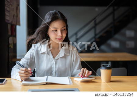 Smart asian woman using calculator holding pen over notebook beside coffee on wooden table in cafe. 140133254