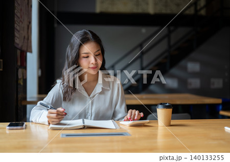 Pretty asian woman using calculator holding pen over notebook beside coffee on wooden table in cafe. Pretty asian woman using calculator holding pen over notebook beside coffee on wooden table in cafe. 140133255