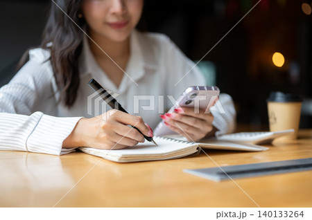 Close up of asian woman looking at phone while writing in notebook beside calculator on wooden table 140133264