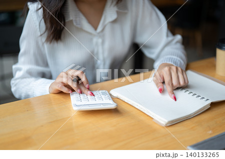 Close up of asian woman looking at phone while writing in notebook beside calculator on wooden table Close up of asian woman looking at phone while writing in notebook beside calculator on wooden table 140133265