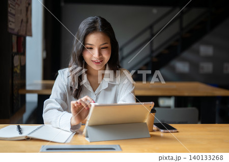 Happy asian woman looking at tablet touching on screen beside notebook sits at wooden table in cafe. Happy asian woman looking at tablet touching on screen beside notebook sits at wooden table in cafe. 140133268