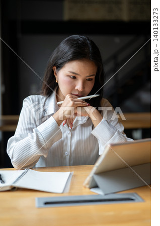 Serious asian woman clasping hands holding pen and looking staring at tablet at wooden table in cafe 140133273