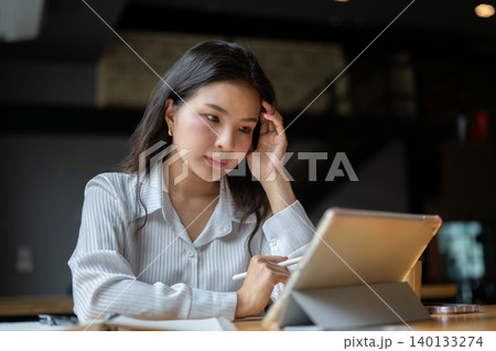 Serious asian woman holding pen touching temple looking at tablet while sits at wooden table in cafe. 140133274