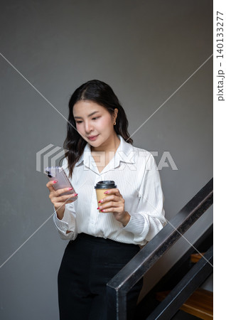 Smart asian woman holding coffee cup looking at phone while standing walking down the stairs in cafe Smart asian woman holding coffee cup looking at phone while standing walking down the stairs in cafe 140133277