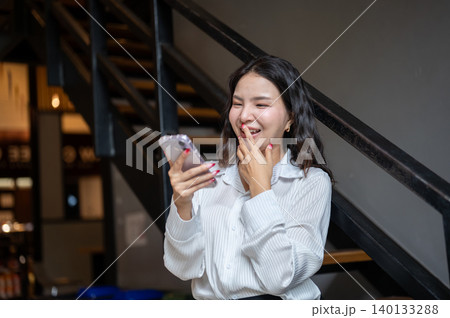 Laughing asian woman covering mouth looking at phone while standing beside stairs in cafe building. Laughing asian woman covering mouth looking at phone while standing beside stairs in cafe building. 140133288