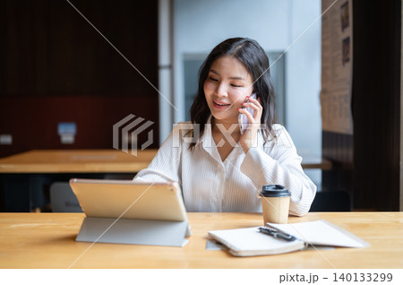 Pretty asian woman talking on phone looking at tablet beside coffee sitting at wooden table in cafe. Pretty asian woman talking on phone looking at tablet beside coffee sitting at wooden table in cafe. 140133299
