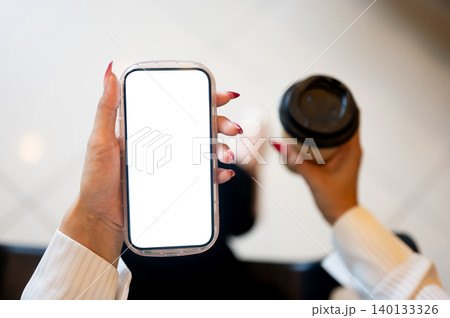 Close up of woman's long nail hands holding blank screen smartphone and coffee while sitting indoors Close up of woman's long nail hands holding blank screen smartphone and coffee while sitting indoors 140133326