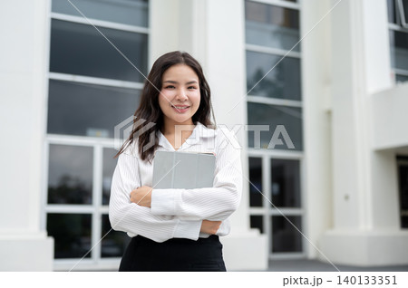 Pretty asian student or businesswoman holding hugging a tablet while standing in front of a building 140133351