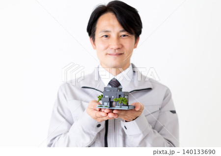 家の住宅模型を持って微笑むグレーの作業着姿のミドル男性ビジネスマン白バック 140133690