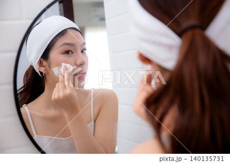 Beautiful young asian woman gently clean on face while looking mirror in bathroom. 140135731