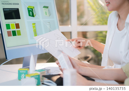 Two asian woman graphic designers analyzing product package prototype, reviewing paper sketch. 140135735