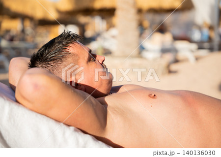 Caucasian man relaxes on a beach lounge chair at a tropical resort under warm sunlight with calm and peaceful expression. Beach vacation and relaxation lifestyle Caucasian man relaxes on a beach lounge chair at a tropical resort under warm sunlight with calm and peaceful expression. Beach vacation and relaxation lifestyle 140136030