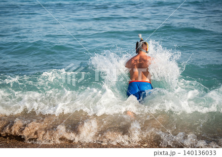 Caucasian man wearing full face snorkeling mask enters sea waves from a sandy beach during active summer vacation by the ocean. Snorkeling and marine adventure 140136033