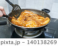 Close-up of marinated pork steaks cooking in savory sauce in a frying pan on a gas stove. A person uses a black spatula to flip the meat. Home cooking and delicious dinner food preparation. 140138269
