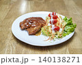 Grilled pork steak with rich gravy sauce served alongside a fresh salad of shredded vegetables, cherry tomatoes, and creamy dressing on a white plate. Gourmet meal on light wooden table background. 140138271