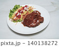 Grilled pork steak glazed with black pepper sauce served on a white plate with fresh vegetable salad featuring carrots, cabbage, and tomatoes on a marble table. Healthy restaurant meal concept. 140138272