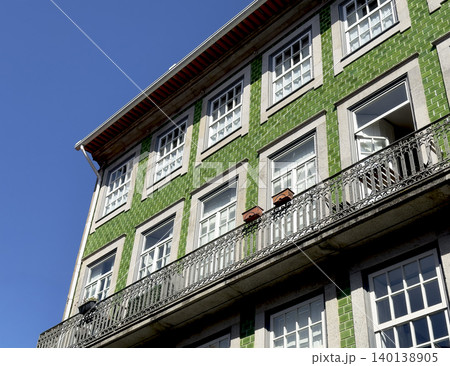Corner view of green azulejo building with balconies under blue sky. Traditional Portuguese facade displays patterned tiles and window details. 140138905
