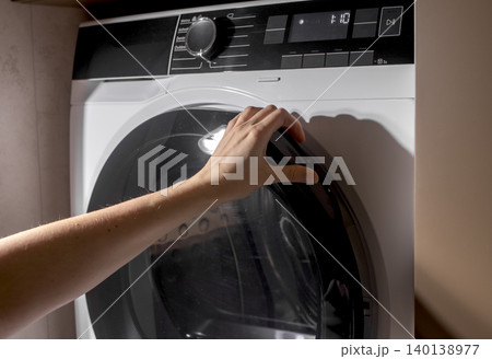 Woman touching drying machine door during cycle. Adult female hand on front loading appliance in laundry room. Domestic washing concept for home, household, lifestyle.. 140138977