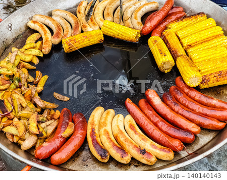 Assorted Grilled Sausages Corn on the Cob and Roasted Potatoes Cooking on Large Outdoor Flat Pan Assorted Grilled Sausages Corn on the Cob and Roasted Potatoes Cooking on Large Outdoor Flat Pan 140140143