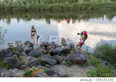 Two women spend a sunny day by the river. One woman poses for photography, while the other squats, capturing serene landscapes and joyful memories. Two women spend a sunny day by the river. One woman poses for photography, while the other squats, capturing serene landscapes and joyful memories. 140141238