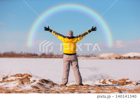 A person wearing a yellow jacket and gray pants stands with arms outstretched, embracing a colorful rainbow arching over a snowy landscape on a clear winter day. 140141340