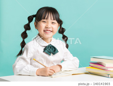 Asian little schoolgirl writing in notebook at desk, focused child studying with books on blue background 140141903