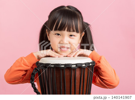 Cute Asian little girl playing drum, smiling child with percussion instrument on pink background, music education concept 140141907