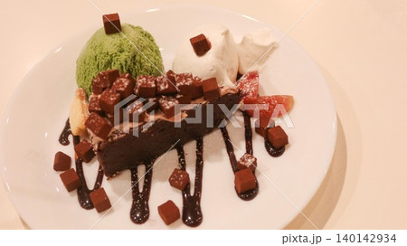 close up of a chocolate brownie cake with green tea matcha ice cream and cream on the sides. Melt chocolate suace and chocolate dice added as decoration on a white plate with copy space 140142934