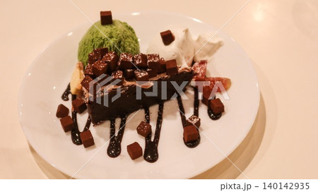 close up of a chocolate brownie cake with green tea matcha ice cream and cream on the sides. Melt chocolate suace and chocolate dice added as decoration on a white plate with copy space close up of a chocolate brownie cake with green tea matcha ice cream and cream on the sides. Melt chocolate suace and chocolate dice added as decoration on a white plate with copy space 140142935