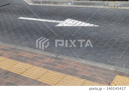 A bold white arrow points right along a paved walkway in a bustling urban area, inviting people to move forward during sunny hours A bold white arrow points right along a paved walkway in a bustling urban area, inviting people to move forward during sunny hours 140145417