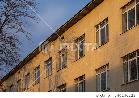 The facade of a beige tiled institutional building is illuminated by golden hour sunlight at dusk, creating long shadows from nearby leafless tree branches on the exterior wall The facade of a beige tiled institutional building is illuminated by golden hour sunlight at dusk, creating long shadows from nearby leafless tree branches on the exterior wall 140146225