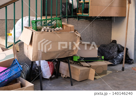 Stacked cardboard boxes and plastic crates piled on a small table outside an urban apartment, representing domestic waste management, recycling preparation, and cluttered living Stacked cardboard boxes and plastic crates piled on a small table outside an urban apartment, representing domestic waste management, recycling preparation, and cluttered living 140146240