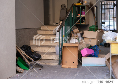 A cluttered staircase entry filled with abandoned cardboard boxes, plastic crates, and household cleaning tools, illustrating urban decay and waste management challenges in a city 140146241