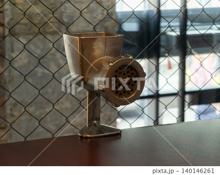 A vintage metal meat grinder rests on a rustic wooden table against a wire mesh background, creating a classic atmosphere ideal for culinary themes or industrial cafe decor A vintage metal meat grinder rests on a rustic wooden table against a wire mesh background, creating a classic atmosphere ideal for culinary themes or industrial cafe decor 140146261