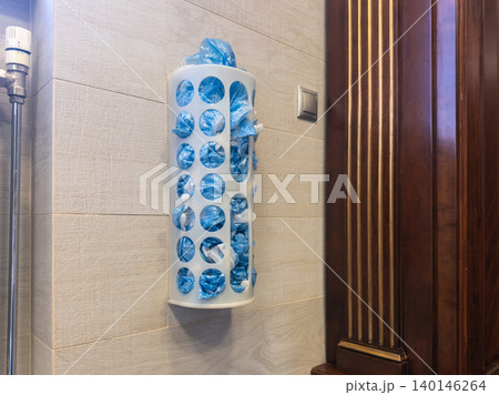 A white plastic wall mounted dispenser filled with blue recycled trash bags in a kitchen, demonstrating organized home storage solutions for waste management and utility items 140146264