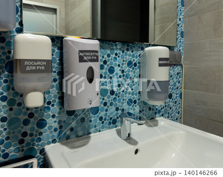 Wall mounted liquid soap and hand sanitizer dispensers above a white ceramic bathroom sink, promoting hygiene and cleanliness in a professional public restroom environment interior 140146266