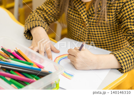 Young girl in a yellow tweed jacket drawing a vibrant rainbow with colorful pencils on paper at a school desk, depicting childhood creativity and artistic learning in a classroom Young girl in a yellow tweed jacket drawing a vibrant rainbow with colorful pencils on paper at a school desk, depicting childhood creativity and artistic learning in a classroom 140146310