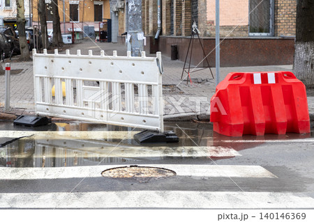 White and red plastic road barriers placed near a puddle on a city street crosswalk to redirect pedestrian traffic safely away from ongoing utility maintenance or repair work zones 140146369