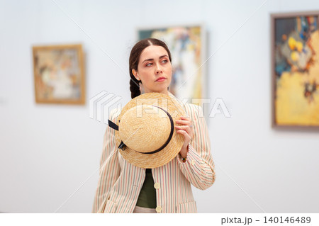 Portrait of a young Caucasian pensive woman elegant jacket and trousers holds straw hat and posing in the art gallery. Painting in background. Concept of cultural education and Museum Day Portrait of a young Caucasian pensive woman elegant jacket and trousers holds straw hat and posing in the art gallery. Painting in background. Concept of cultural education and Museum Day 140146489
