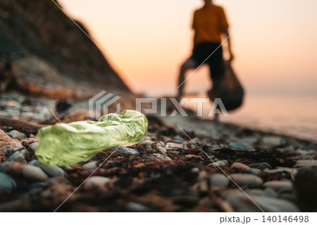 Close up of dirty green plastic bottle lies on the beach. Defocused silhouette of a person holding a plastic bag with garbage on the background. The concept of envirnmental pollution Close up of dirty green plastic bottle lies on the beach. Defocused silhouette of a person holding a plastic bag with garbage on the background. The concept of envirnmental pollution 140146498