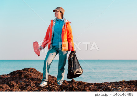 A young Caucasian female volunteer in a jacket poses with a large garbage bag and a plastic bottle in her hands. In the background, the sky and the sea. Copy space A young Caucasian female volunteer in a jacket poses with a large garbage bag and a plastic bottle in her hands. In the background, the sky and the sea. Copy space 140146499