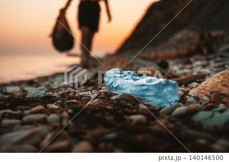Close up of dirty blue plastic bottle lying on the beach. Defocused silhouette of a person holding a plastic bag with garbage on the background. The concept of Earth Day Close up of dirty blue plastic bottle lying on the beach. Defocused silhouette of a person holding a plastic bag with garbage on the background. The concept of Earth Day 140146500